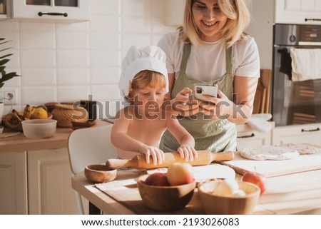 Pretty young woman in chef's apron and her little son are preparing apple pie at home in cozy kitchen. Kid in cute chef's hat. She takes pictures of the process of making cake