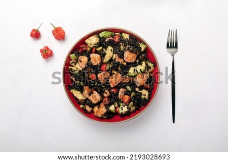 A red bowl of black rice with baked salmon, avocado, tomatoes, chili peppers and edamame on a white marble background. 