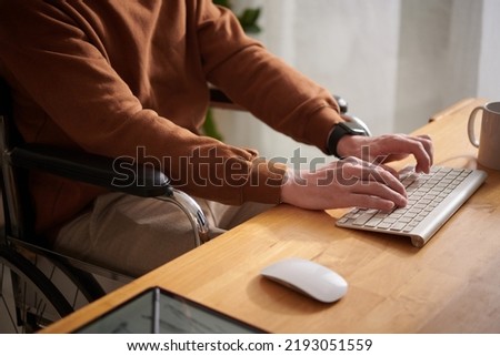 Closeup image of young man sitting in wheelchair working on laptop at his desk
