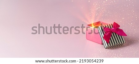 Open pink box with black stipes tap and red bow on pink background with golden spakling and light. Banner with copy space