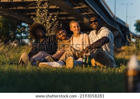 Happy diverse group of young friends sitting together on a blanket on the grass, drinking beer, telling stories, laughing, and enjoying the evening during their van camping trip. High quality photo