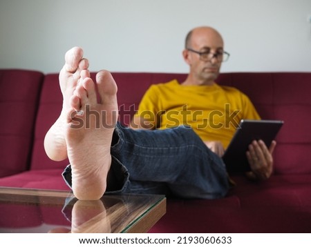 Man sitting on a red sofa with feet up on coffee table in living room, reading from a tablet computer wearing casual clothes and glasses. Feet and table are in sharp focus, man is soft focus. 