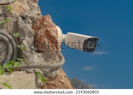 Close-up of a white CCTV camera attached to a rock near a private house against a blue sky. Tools for regulating the safety of people and private territory.