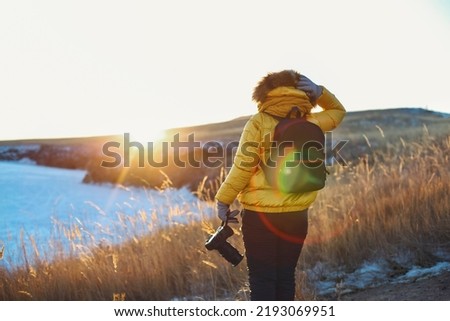 Female traveller looking to Baikal lake in winter time. Woman holding professional camera in her hand wearing bright yellow winter clothes n sunset. Ogoy Island, Baikal Lake, Russia