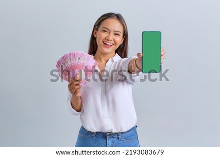 Cheerful young Asian woman showing blank screen mobile phone and holding money banknotes isolated over white background