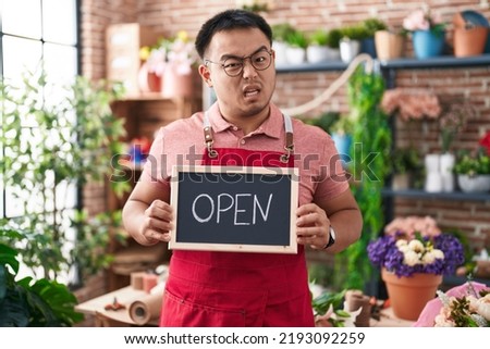 Chinese young man working at florist holding open sign clueless and confused expression. doubt concept. 