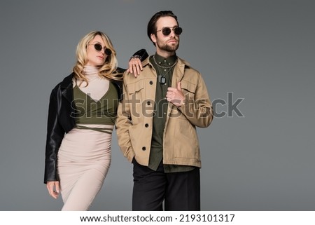 stylish man in sunglasses posing with hand in pocket near blonde woman in leather jacket and dress isolated on grey Royalty-Free Stock Photo #2193101317