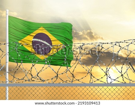 Flagpole with the flag of Brazil against the sky and behind a fence with barbed wire. The concept of protecting the borders of territories.