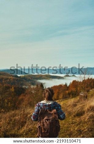 Rear view woman hiker backpacker wearing a checkered jacket enjoying beautiful view, walks on hills over mountain summit at sunrise