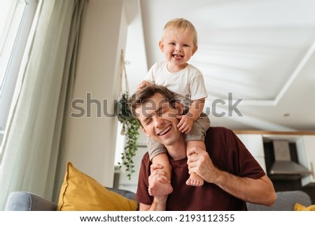 Laughing father giving his kid piggyback ride on shoulders, baby boy pulling daddy's hair, father and son spending time together on sunday morning in living-room. Dad playing with his toddler boy Royalty-Free Stock Photo #2193112355