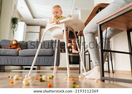 Side view of cute caucasian baby sitting in high chair with piece of vegetable looking with interest on messy and dirty floor covered with broccoli, carrot and cauliflower. Baby feeding concept Royalty-Free Stock Photo #2193112369