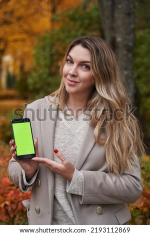 portrait of a young beautiful woman with long blond hair in autumn against the background of trees with yellow leaves holds a smartphone with a green screen in her hands