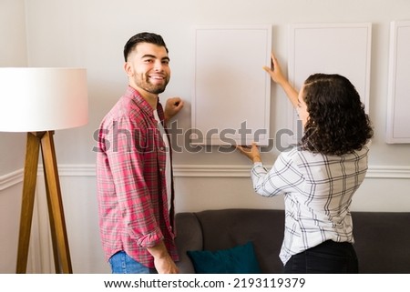 Attractive young man smiling while making eye contact and putting new paintings in the wall with his partner at home 