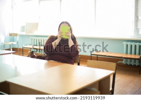 Teacher hides her face behind book. Teacher at table. Funny picture of teacher. Teacher in Russia.