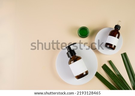 mock-up of brown cosmetics bottle with dispenser and white label, bottle with pipette on white round podium on beige colored background, green palm leaf, top view