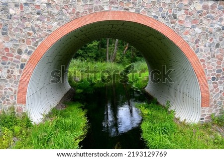 A view of a stone and brick bridge with an arch inside of it used to transport water with both banks of the water reservoir covered with grass, herbs, and other plants seen in Poland in summer
