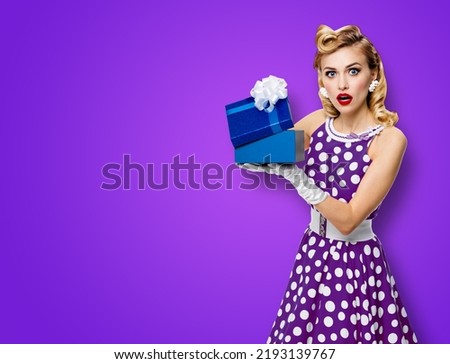 Image of beautiful woman dressed in pin up dress with polka dot white gloves opening gift box, isolated on violet purple background. Blond pinup model in retro fashion studio concept. Big sales deals.