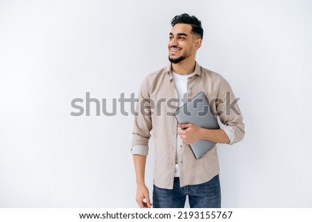 Positive friendly modern arabian or indian young man with beard, wearing casual shirt, standing on isolated white background, holding laptop, looking to the side, smiling, dreaming, thinking Royalty-Free Stock Photo #2193155767