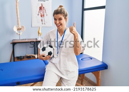 Young woman working at football therapy clinic smiling happy and positive, thumb up doing excellent and approval sign 