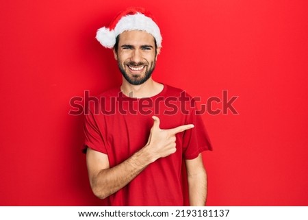 Young hispanic man wearing christmas hat cheerful with a smile of face pointing with hand and finger up to the side with happy and natural expression on face 
