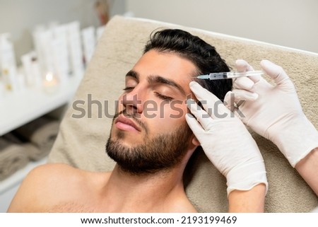 From above anonymous beautician in latex gloves injecting botulinum toxin into forehead of bearded young male client during cosmetology session in beauty studio Royalty-Free Stock Photo #2193199469