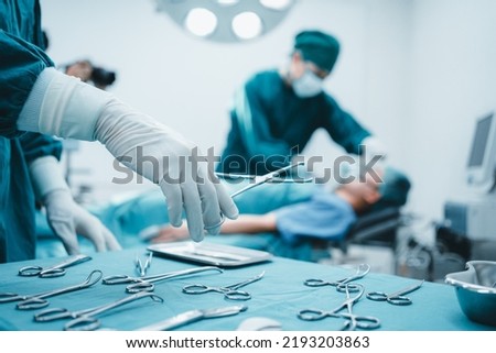 Close up surgeon doctor's hand with hygiene glove taking sterile surgical instrument tool, equipment in operating room patient in background for surgery. Hospital, medical healthcare emergency concept