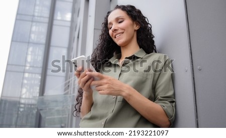 Confident 30s adult woman success businesswoman smiling texting in mobile app using phone online e-commerce service in internet chatting with smartphone outdoors leaning to wall of office building
