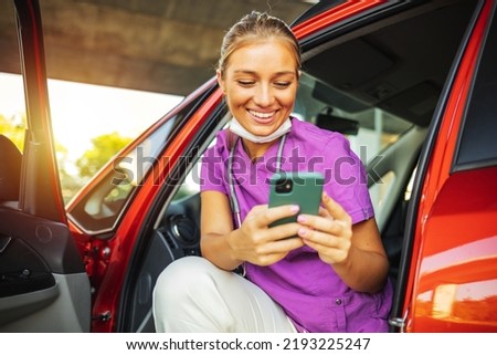 A smiling young nurse dressed in her scrubs uniform sitting in her car holding her mobile phone, taking a break from work.