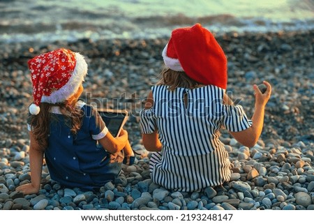 two sister girls in santa claus hats are sitting on the seashore at sunset, in the hands of one girl is a tablet 