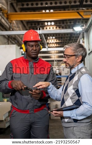 Quality Control Inspector With Digital Tablet Talking To African American CNC Machine Operator. Multiracial Industrial Co-Workers Standing And Talking About Production Process.