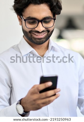 Happy indian young business man, company worker, professional employee, stock market trader holding smartphone using cell phone mobile apps checking financial data in app at work in office. Vertical
