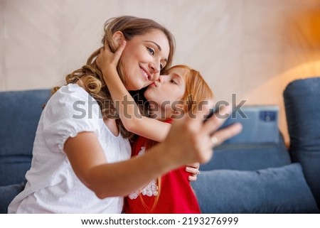 Beautiful mother and daughter having fun spending leisure time together at home, taking selfies using smart phone, little girl kissing her mom
