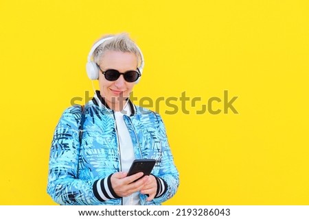 Middle Aged Woman, Short HairStyle, Casual Look With White Headphones On Yellow Background. Listening Music or Podcast. Using Smartphone and Smiling, Older Adults and Technology Use