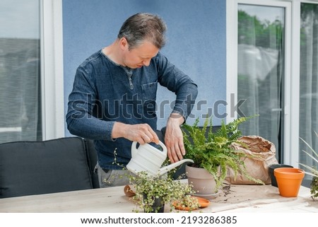 A young man transplants homemade flowers into pots. A gardener is watering a fern from a watering can on the veranda of his house