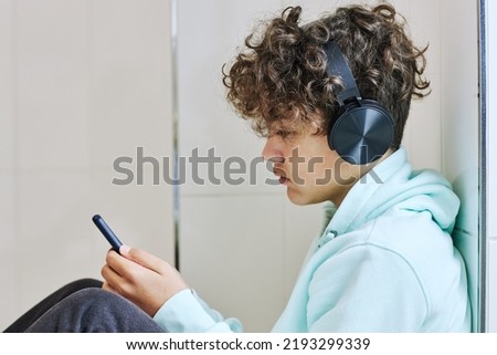Minimal side view portrait of teen schoolboy sitting on floor and using smartphone with headphones Royalty-Free Stock Photo #2193299339