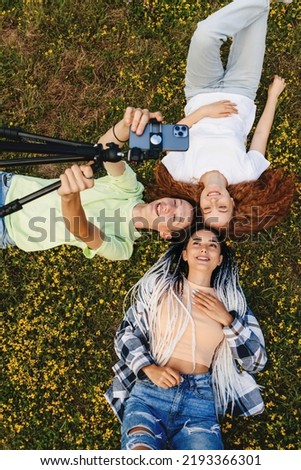 Three teenagers lying on the grass in the park making videos to post on the internet. Young and influential. Modern communication, social media and gadgets.