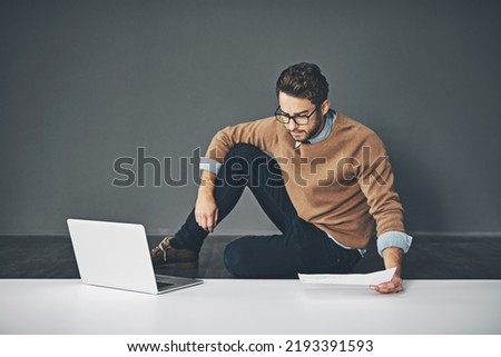 Business man working on a laptop, reading a report and going through finance paperwork while sitting on the floor in an office at work. Serious professional manager looking at papers with copyspace