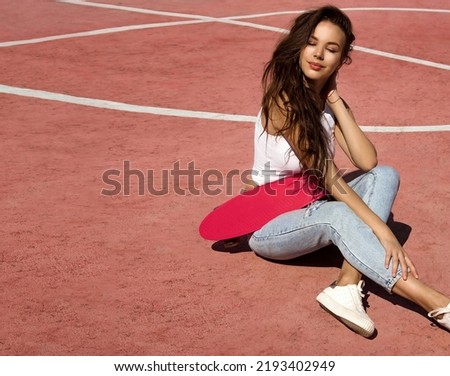 teen leisure. young fit girl with long legs in sports top and jeans sits casual with pink skateboard on red court background sports ground at sunny day and smiles. lifestyle sport concept, free space