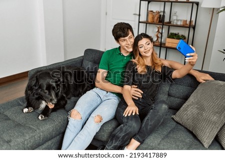 Young caucasian couple making selfie by the smartphone sitting on the sofa with dog at home.
