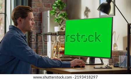 Young Handsome Man Working from Home on Desktop Computer with Green Screen Mock Up Display. Male Checking Corporate Accounts, Messaging Colleagues. Loft Living Room with Big Window.