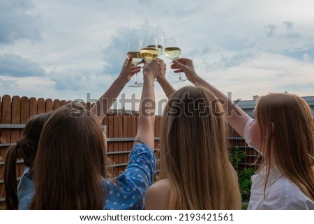 Back view of group of young women with long hair raising clinking glasses with white wine champagne on cloudy sky background celebrating in backyard. Summer, celebration, holiday, hen-party