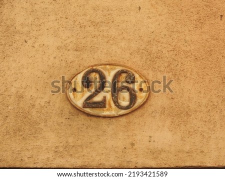 House number (address) twenty six (26) sign, black numbered white plate (closeup) against cream and off white wall of old building background. Grunge, texture, wallpaper   