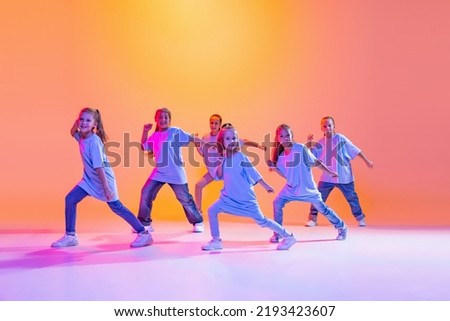 Hip-hop dance, street style. Happy children, little active girls in casual style clothes dancing isolated on orange background in purple neon light. Concept of music, fashion, art