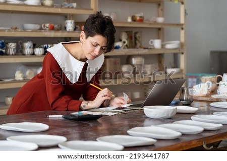 Young woman talented master potter run pottery business, focused busty female entrepreneur calculating cost to produce ceramic items. Small business and entrepreneurship in art, self-employment tax Royalty-Free Stock Photo #2193441787