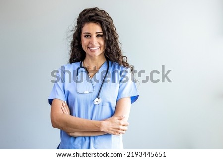 Modern Medical Education Concept. Portrait Of Smiling Female Doctor In Blue Coat Posing With Folded Arms Over Light Background. Portrait Of Female Nurse Wearing Scrubs In Hospital