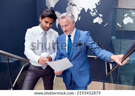 Male Colleagues Meeting On Stairs Of Office Discussing Document With World Map In Background