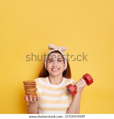 Portrait of smiling woman wearing striped shirt and hair band holding red dumbbell and cookies, posing isolated over yellow background, looking up at copy space for advertisement or promotion.