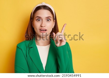 Portrait of astonished beautiful woman wearing green jacket and hair band posing isolated over yellow background, pointing away at copy space, indicating advertisement area for your goods,