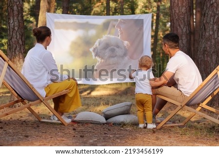 Portrait of happy family sitting in the forest on deck chairs with their little daughter and watching movie on projector, parents playing with their cute kid and enjoying nature and fresh air.