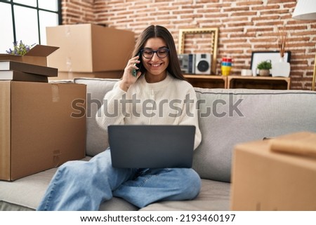 Young hispanic woman sitting on the sofa at new home using laptop peeking in shock covering face and eyes with hand, looking through fingers afraid 